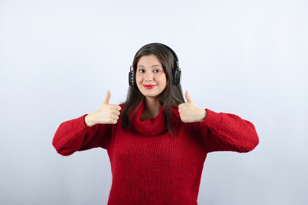 Modello di giovane donna in maglione rosso con le cuffie che mostra i pollici in su