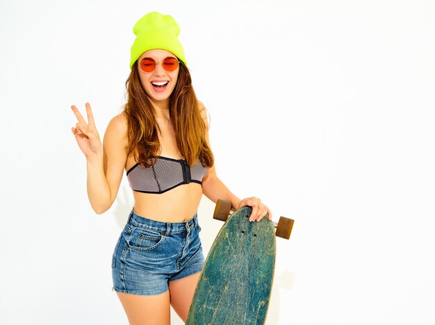 Modello di giovane donna sorridente alla moda in costumi da bagno casual estate e berretto giallo in posa con scrivania longboard, mostrando il segno di pace. Isolato su bianco