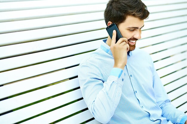 Modello di giovane uomo d'affari sorridente bello seduto sulla panchina del parco in panno casual hipster parlando sul telefono cellulare