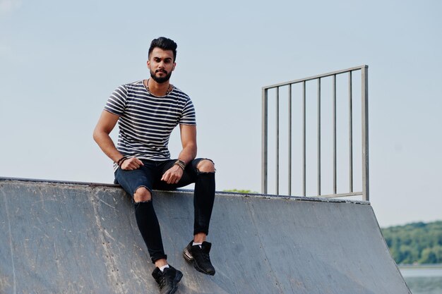 Modello di uomo bello e alto con barba araba con camicia spogliata posata all'aperto allo skate park Ragazzo arabo alla moda