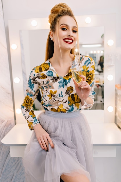 Modello elegante attraente in gonna di tulle, con coiffure di lusso, bel trucco che sorride alla macchina fotografica nel salone di parrucchiere con un bicchiere di champagne