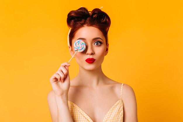 Modello femminile adorabile che tiene caramella dura. Studio shot di ispirata ragazza allo zenzero con lecca-lecca isolato su spazio giallo.