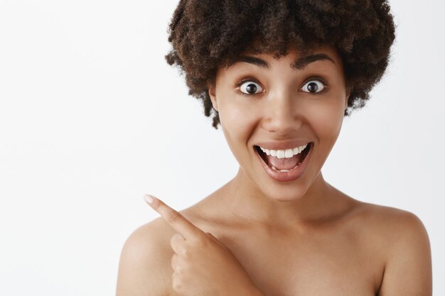 Modello femminile afroamericano divertito ed elettrizzato con taglio di capelli ricci in piedi nudo con la pelle pulita, che punta all'angolo in alto a sinistra, sorridendo con gioia, essendo colpito e soddisfatto del prodotto