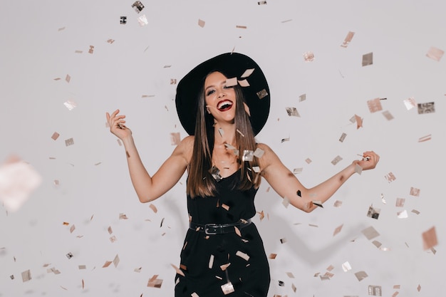 Modello femminile alla moda attraente in costume della strega che prepara per la festa di Halloween sulla parete isolata con i coriandoli che ballano, divertendosi, sorridente. Compleanno, vacanza