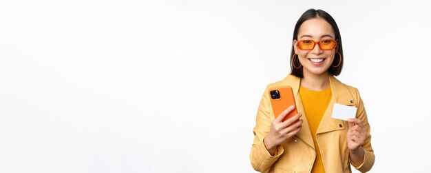 Modello femminile asiatico alla moda di acquisto online in occhiali da sole che tengono carta di credito e telefono cellulare smilin