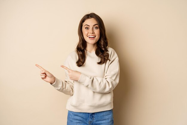 Modello femminile attraente allegro che punta il dito a sinistra, mostrando pubblicità del negozio, banner o logo, in piedi in maglione su sfondo beige