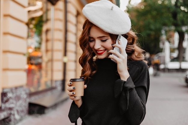 Modello femminile carino con capelli ondulati di zenzero parlando al telefono. Signora francese di buon umore in posa con lo smartphone sulla strada.