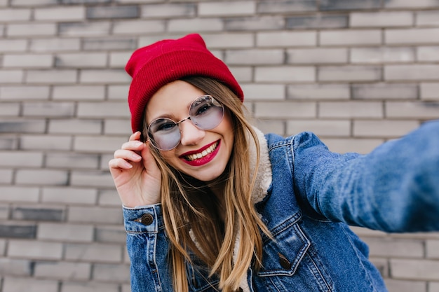 Modello femminile caucasico splendido che tocca scherzosamente i suoi occhiali da sole. Colpo esterno di donna alla moda che ride con i capelli chiari in posa vicino al muro di mattoni.