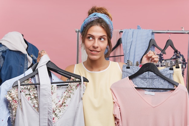 Modello femminile confuso che tiene i ganci con vestiti con entrambe le mani, cercando di scegliere qualcosa di adatto. Femmina che ha esitazione tra gli acquisti. Concetto di persone, moda, vendita, abbigliamento e shopping