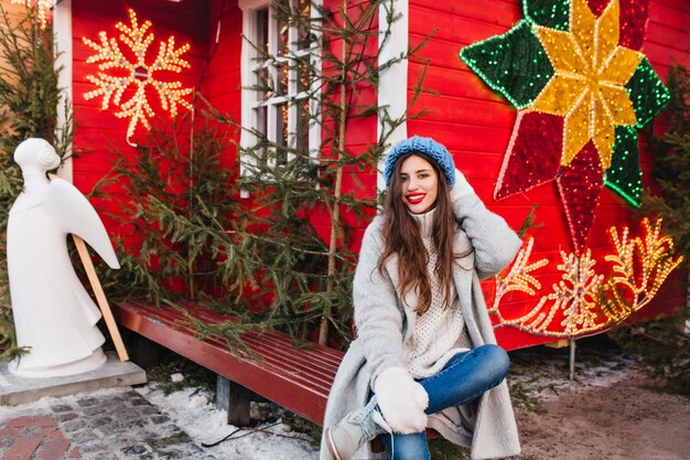 Modello femminile dai capelli lunghi si siede sulla panca di legno vicino alla casa rossa decorata per Natale. Attraente ragazza bruna in posa dopo le vacanze di Capodanno accanto a verdi alberi e sculture di angeli.