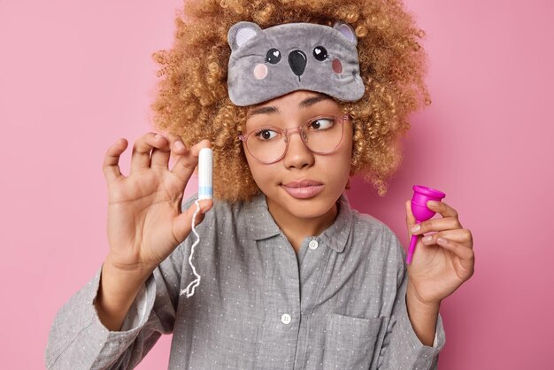 Modello femminile dai capelli ricci esitante tiene diversi tipi di prodotti per l'igiene femminile sceglie tra tampone e coppetta mestruale vestita in pigiama posa su sfondo rosa