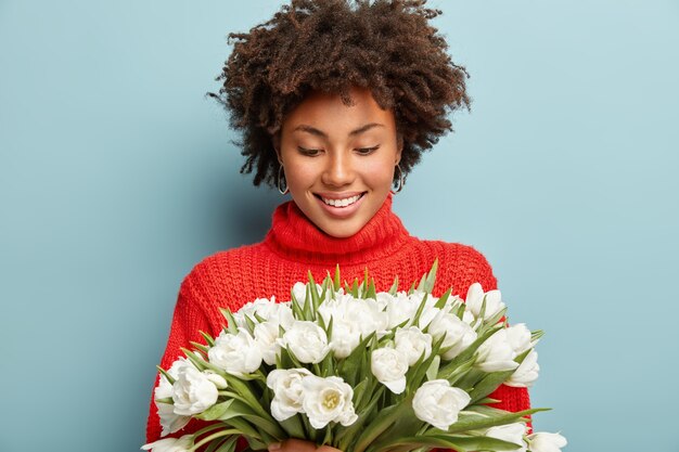 Modello femminile dall'aspetto piacevole soddisfatto con i capelli ricci, guarda felicemente sui fiori primaverili bianchi