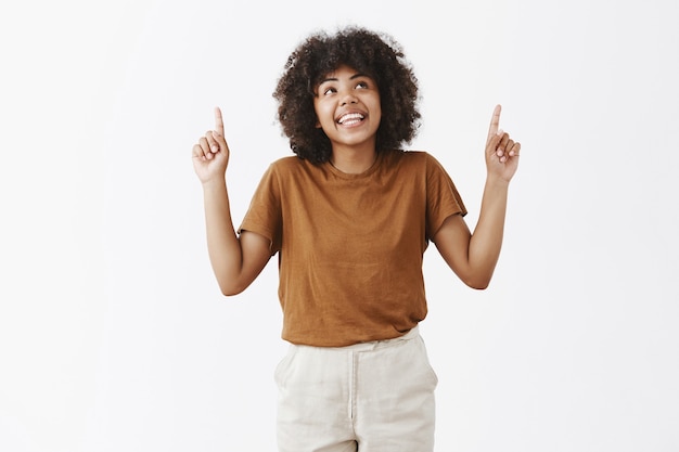 modello femminile dalla pelle scura attraente sognante e creativo con acconciatura afro in abito alla moda guardando e rivolto verso l'alto con un sorriso divertito gioioso guardando lo spazio della copia