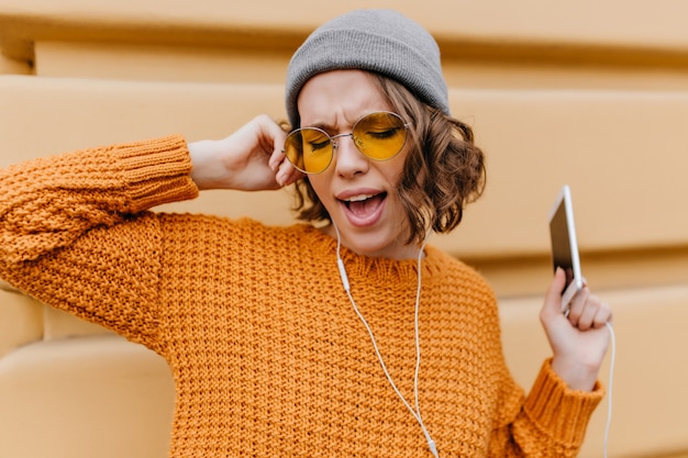 Modello femminile divertente con i capelli ricci corti che canta la canzone preferita mentre si trova sulla strada con lo smartphone