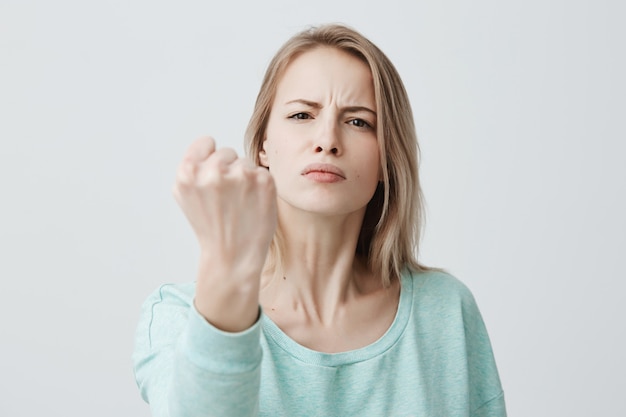 Modello femminile europeo feroce e fiducioso con i capelli lunghi biondi, mostra il pugno chiuso davanti a sé come se fosse pronto per la lotta o qualsiasi sfida, inseguendo le labbra, ha determinato l'espressione del viso