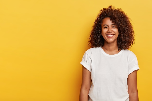 Modello femminile rilassato con acconciatura afro riccia, sorride dalla felicità, felice di avere una giornata di successo, guarda dritto, indossa una maglietta bianca, ride al coperto sopra il muro giallo, copia spazio