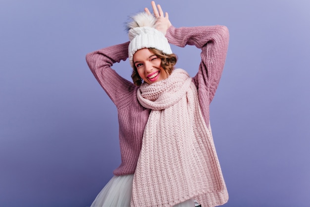 Modello femminile soddisfatto con l'espressione del viso felice in posa in abiti invernali e sorridente. Donna dai capelli corti in sciarpa che esprime emozioni positive sulla parete viola.