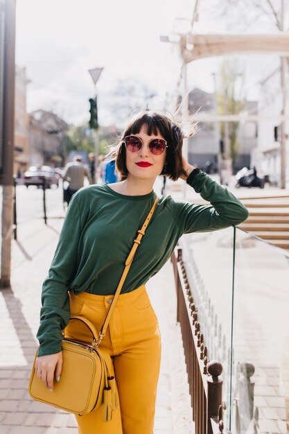 Modello femminile sveglio in pantaloni gialli che tocca i suoi capelli con il sorriso. Ritratto all'aperto di incantevole ragazza bruna in occhiali da sole in posa con borsa elegante.