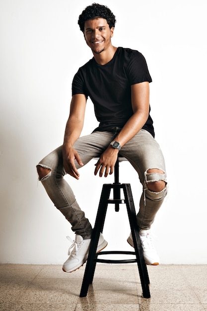 Modello giovane atletico sorridente rilassato in jeans strappati, scarpe da ginnastica bianche e t-shirt nera semplice su sgabello da bar con pareti bianche