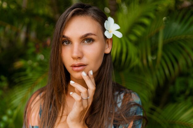 Modello grazioso vicino della donna con il fiore di plumeria in capelli che posano nella natura tropicale.