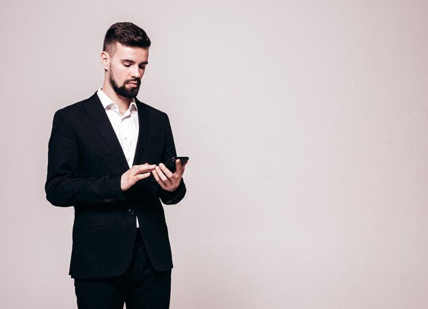 Modello lambersexual bello fiducioso elegante hipster Uomo moderno sexy vestito con un elegante abito nero Moda maschile in posa in studio Holding smartphone Guardando lo schermo del cellulare Utilizzando le app