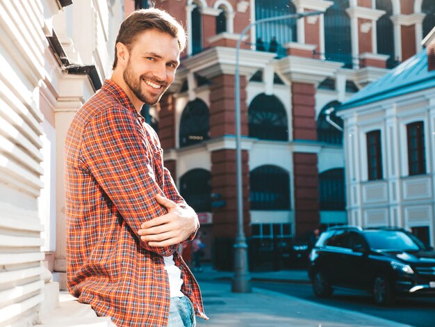 Modello lambersexual sorridente bello dei pantaloni a vita bassa alla moda. L'uomo moderno si è vestito in camicia a scacchi rossa. Moda maschile in posa vicino al grattacielo sullo sfondo della strada. All'aperto al tramonto