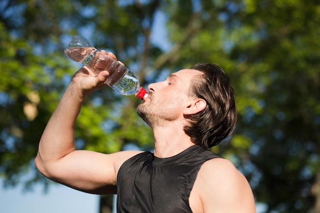 Modello sportivo di formazione etnica caucasica all'aperto. Uomo stanco che beve acqua da una bottiglia di plastica dopo il tempo di fitness e si esercita nel parco cittadino in una bella giornata estiva.