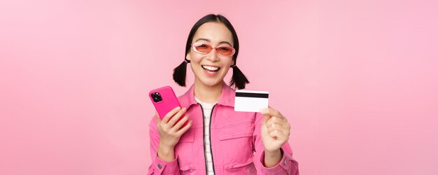 Moderna bella ragazza asiatica che ride e sorride con la carta di credito del telefono cellulare che acquista online pagando con lo smartphone in piedi su sfondo rosa