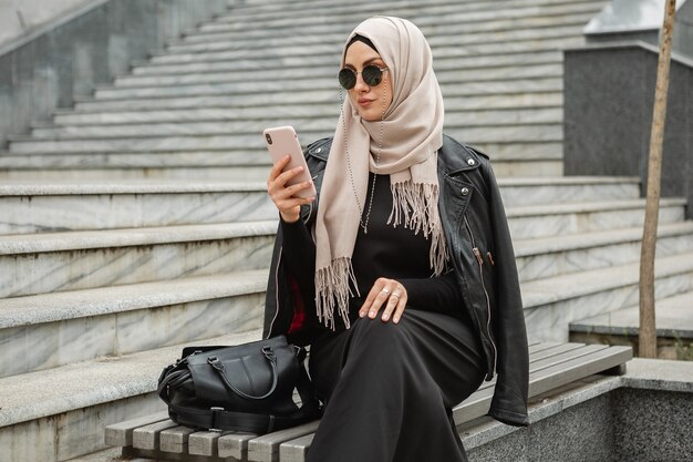 Moderna ed elegante donna musulmana in hijab, giacca di pelle e abaya nera che cammina in una strada cittadina usando lo smartphone