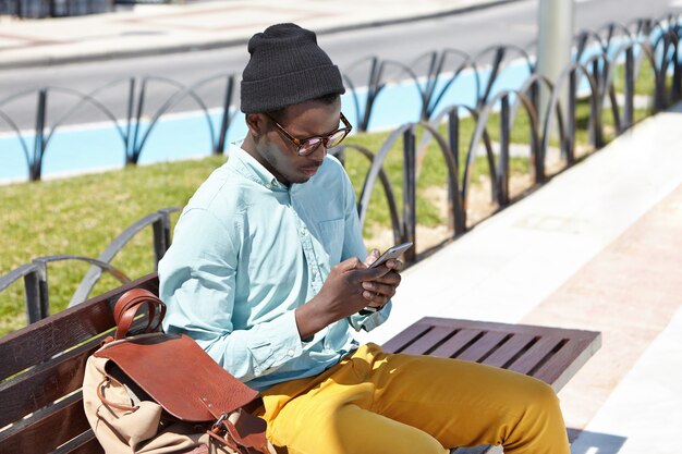 Moderna giovane hipster dalla pelle scura in elegante copricapo e occhiali da sole usando la città libera wi-fi su gadget elettronici all'aperto, seduto su una panchina di legno nel parco in attesa di amici prima di camminare