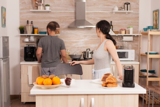 Moglie allegra che parla con il marito in cucina mentre tosta il pane per la colazione. Giovane coppia al mattino che prepara un pasto insieme con affetto e amore