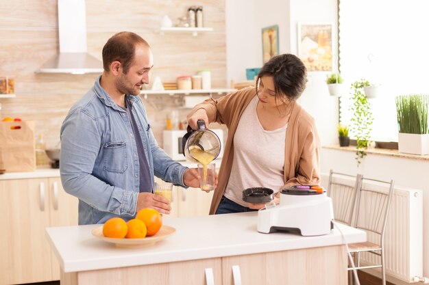 Moglie versando un gustoso frullato mentre il marito tiene il bicchiere. Stile di vita sano e spensierato, mangiare dieta e preparare la colazione in un'accogliente mattinata di sole