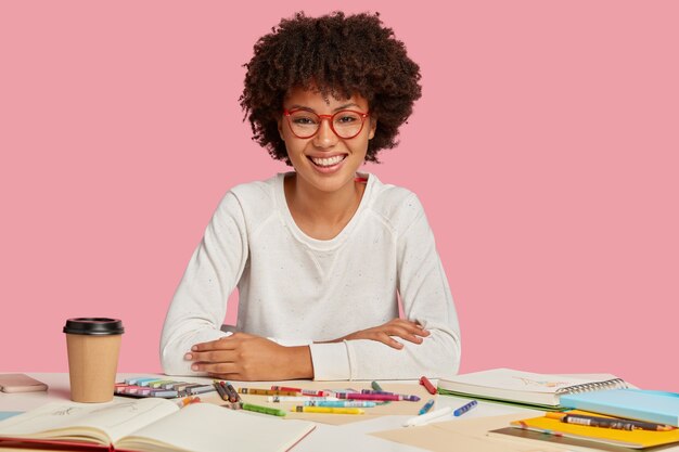 Moltiplicatore femminile positivo con taglio di capelli afro, sorriso piacevole sul viso, ama il suo lavoro, ha un vero talento per fare illustrazioni, gode di bevande calde, isolato su un muro rosa. Concetto di creatività