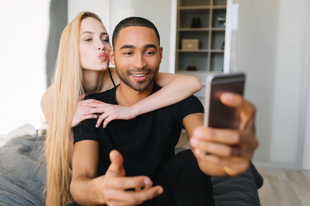 Momenti di coppia incantevole di bel ragazzo e giovane donna graziosa con lunghi capelli biondi che si divertono, facendo selfie sul telefono. Rilassarsi sul letto a casa in un appartamento moderno, baciarsi, innamorarsi