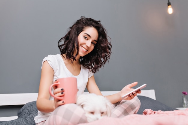 Momenti dolci felici di giovane bella donna con capelli ricci castana tagliati in pigiama agghiacciante sul letto con il cane in appartamento moderno. Sorridere, navigare in internet, rilassarsi nell'intimità di casa