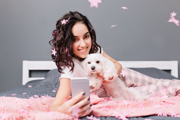 Momenti dolci felici di giovane bella donna in pigiama con capelli ricci castani tagliati facendo selfie foto con il cane in orpelli rosa sul letto in appartamento moderno. Sorridere, esprimere positività