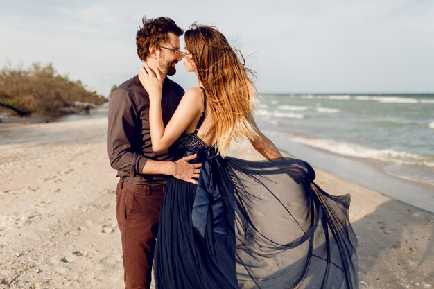 Momenti romantici di bella coppia, donna alla moda e uomo in posa all'aperto vicino al mare. Incredibile vestito blu e abbigliamento casual. Vacanza in luna di miele.