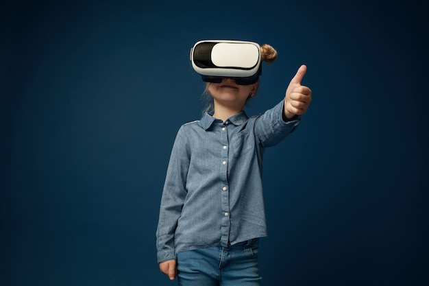 Mostra la tua mente. Bambina o bambino in jeans e camicia con occhiali per cuffie da realtà virtuale isolati su sfondo blu studio. Concetto di tecnologia all'avanguardia, videogiochi, innovazione.