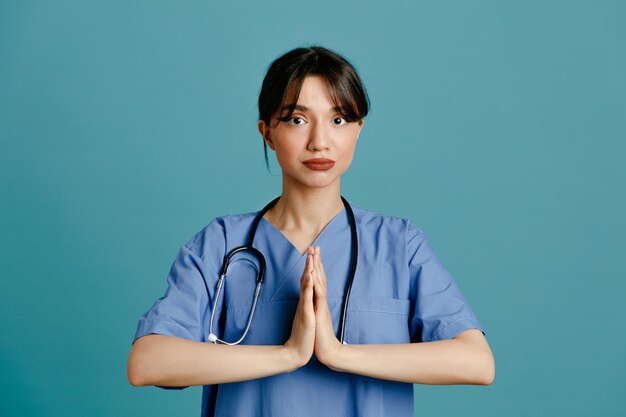 Mostrando il gesto di preghiera giovane dottoressa che indossa uno stetoscopio fitth uniforme isolato su sfondo blu