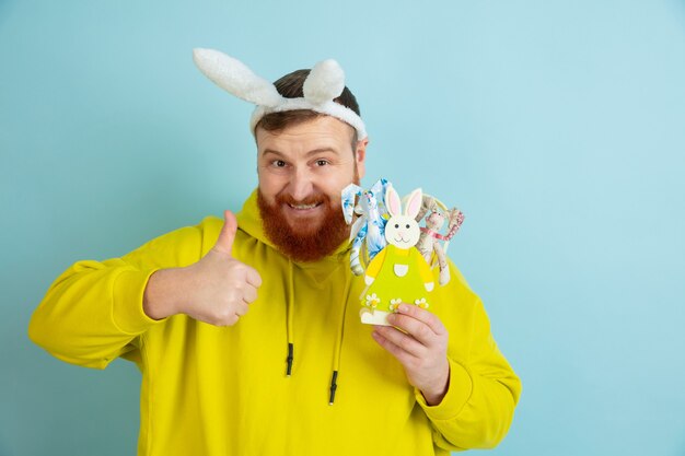 Mostrando il pollice in su. Uomo caucasico come un coniglietto di Pasqua con abiti casual luminosi su sfondo blu studio. Auguri di buona Pasqua. Concetto di emozioni umane, espressione facciale, vacanze. Copyspace.