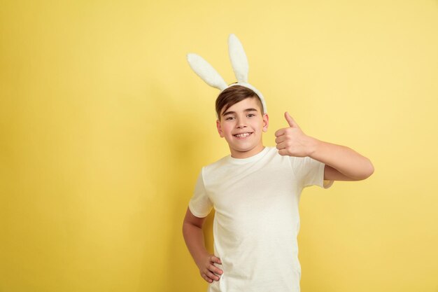 Mostrando pollice in su. Ragazzo caucasico come un coniglietto di Pasqua su sfondo giallo studio. Auguri di buona Pasqua.