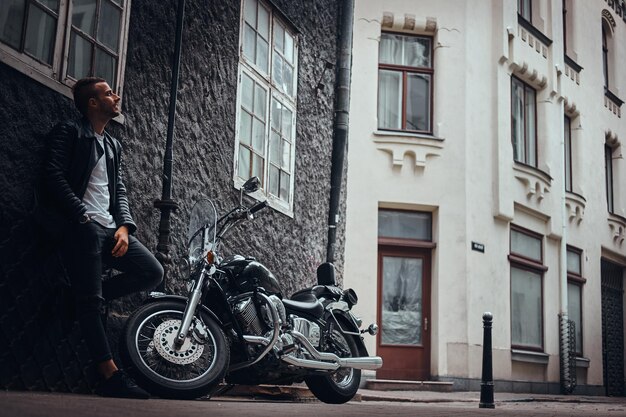 Motociclista alla moda vestito con una giacca di pelle nera e jeans appoggiato a un muro vicino alla sua moto retrò su una strada della vecchia Europa.