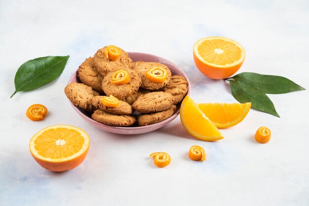 Mucchio fatto in casa di biscotti in una ciotola rosa e fette di arancia o tagliate a metà su superficie bianca.