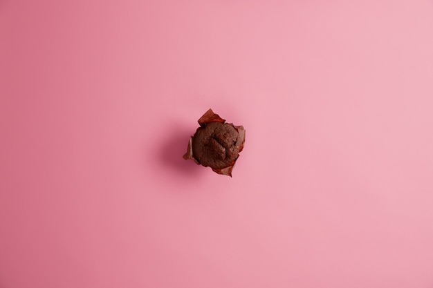 Muffin al cioccolato dolce delizioso appena sfornato in carta marrone fotografato dall'alto, isolato su sfondo rosa, pronto da mangiare. Cibo spazzatura, dessert, calorie e concetto di nutrizione. Messa a fuoco selettiva