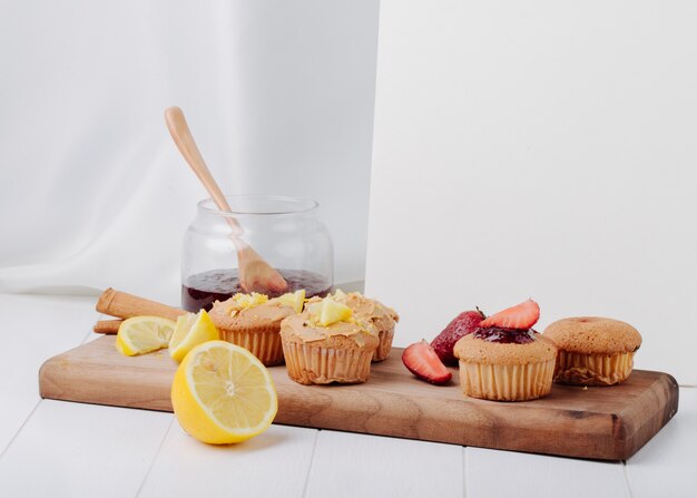 Muffin di vista laterale con le fragole e il limone su una lavagna con un barattolo di inceppamento e un taccuino bianco