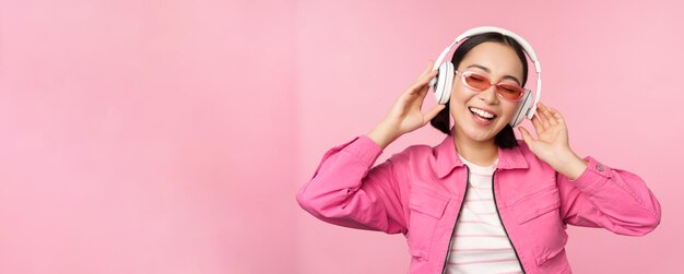Musica d'ascolto della ragazza asiatica alla moda di Dancing in cuffie che posano contro il fondo rosa