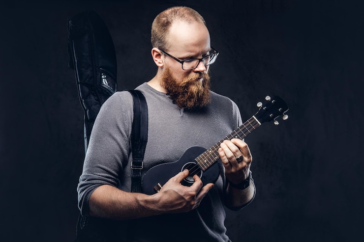 Foto gratuito musicista maschio barbuto dai capelli rossi che indossa occhiali vestito con una maglietta grigia che suona su un ukulele. isolato su sfondo scuro con texture.