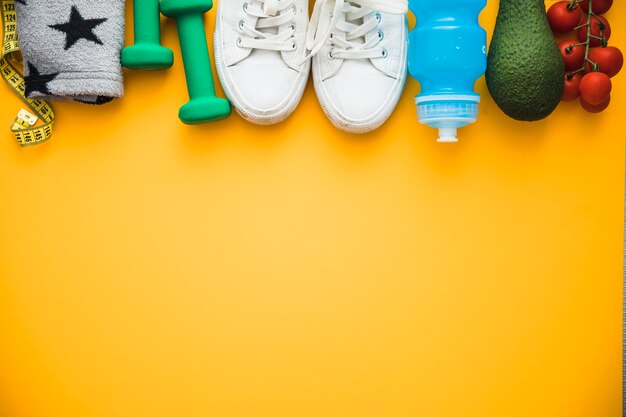 Nastro di misurazione; fascia da braccio; manubri; scarpe; bottiglia d&#39;acqua avocado e pomodorini su sfondo giallo
