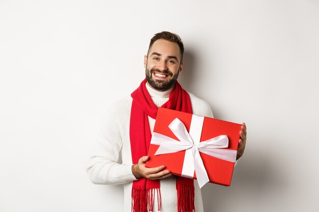 Natale. Bel giovane che tiene in mano un regalo e sorride, augurando buone feste e facendo regali, in piedi su sfondo bianco