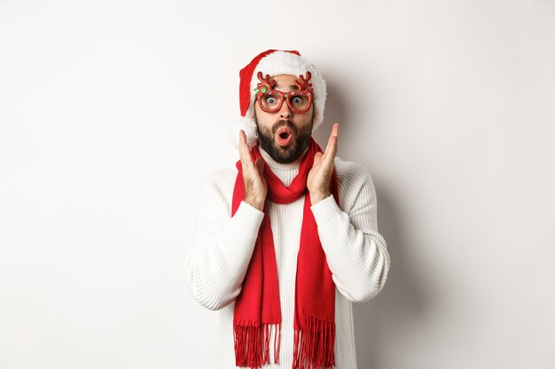 Natale, Capodanno e concetto di celebrazione. L'uomo in occhiali da festa e cappello da Babbo Natale sembra sorpreso, ascolta l'offerta di shopping, in piedi su sfondo bianco.